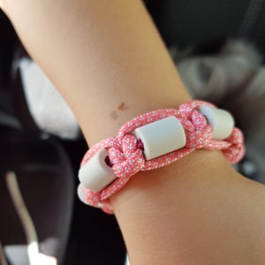 Könnte beinhalten: Ein geflochtenes Armband in Pink und Weiß mit weißen Perlen. Das Armband wird am Handgelenk getragen.