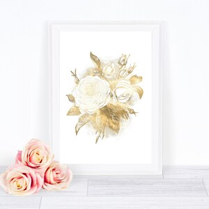 May include: A framed floral art print featuring a watercolor illustration of white roses and gold leaves. The artwork is displayed in a white frame. Three pink roses are in the foreground, adding a touch of color to the composition.