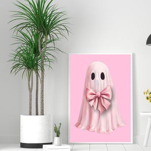 Op de afbeelding: Een ingelijste print met een grillige spookillustratie. De geest is afgebeeld in een zachte roze tint, versierd met een grote bijpassende strik. De achtergrond is effen roze en het kunstwerk wordt gepresenteerd in een witte lijst.