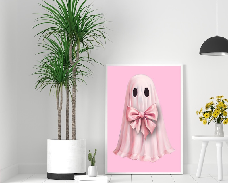 Op de afbeelding: Een ingelijste print met een grillige spookillustratie. De geest is afgebeeld in een zachte roze tint, versierd met een grote bijpassende strik. De achtergrond is effen roze en het kunstwerk wordt gepresenteerd in een witte lijst.