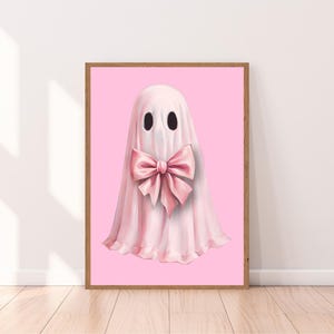 Op de afbeelding: Een ingelijste print met een illustratie van een roze spook met een grote bijpassende strik op een roze achtergrond. De lijst van natuurlijk hout voegt warmte toe. De afbeelding heeft een speelse en charmante uitstraling, geschikt voor een kinderkamer of een eigenzinnige inrichting.
