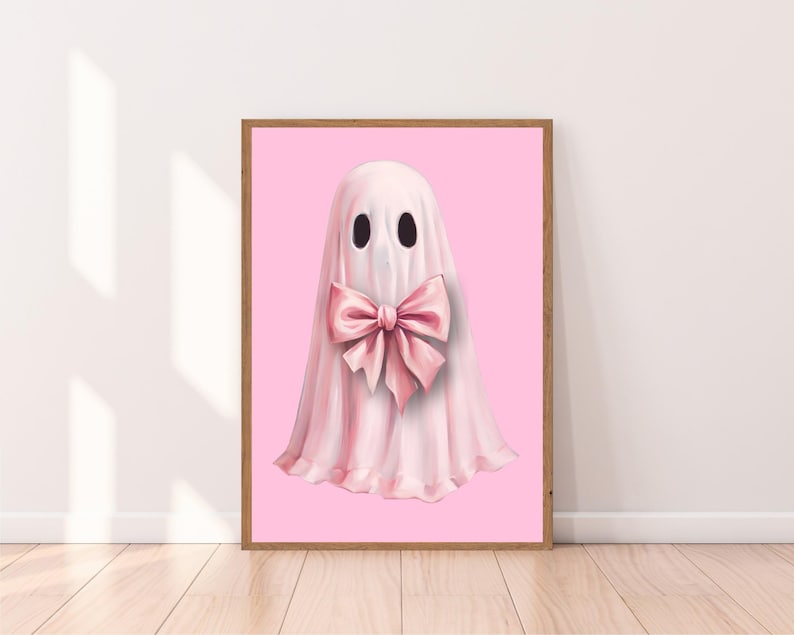 Op de afbeelding: Een ingelijste print met een illustratie van een roze spook met een grote bijpassende strik op een roze achtergrond. De lijst van natuurlijk hout voegt warmte toe. De afbeelding heeft een speelse en charmante uitstraling, geschikt voor een kinderkamer of een eigenzinnige inrichting.