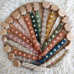 Op de afbeelding: Een verzameling van 18 houten baby-bijtjes met verschillende dierenontwerpen. Elke bijtje is bevestigd aan een macramékoord met verschillende kleuren en kralen. De koorden zijn geweven met verschillende letterkralen, die namen spellen.