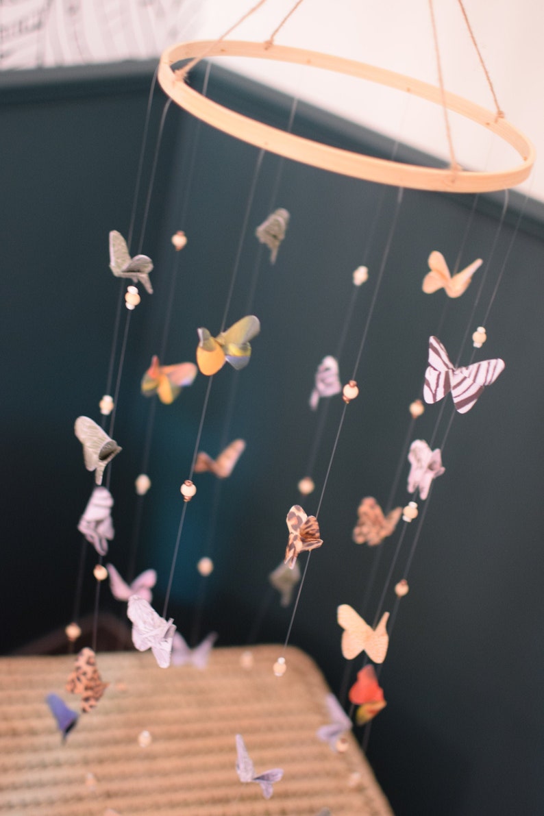 Puede incluir: Un m&oacute;vil de anillo de madera con recortes de mariposas de colores que cuelgan de &eacute;l. Las mariposas est&aacute;n hechas de papel y tienen diferentes patrones y colores. El m&oacute;vil est&aacute; colgado del techo y est&aacute; rodeado por una pared azul oscuro.