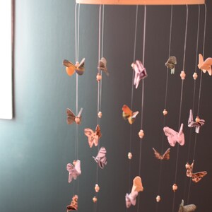 Puede incluir: Un m&oacute;vil de madera con un marco circular y m&uacute;ltiples cuerdas que cuelgan. Cada cuerda tiene un peque&ntilde;o adorno de papel en forma de mariposa de colores. Las mariposas est&aacute;n hechas de diferentes patrones y colores, incluyendo rosa, azul, verde y amarillo. El m&oacute;vil est&aacute; colgado del techo contra una pared verde oscuro.