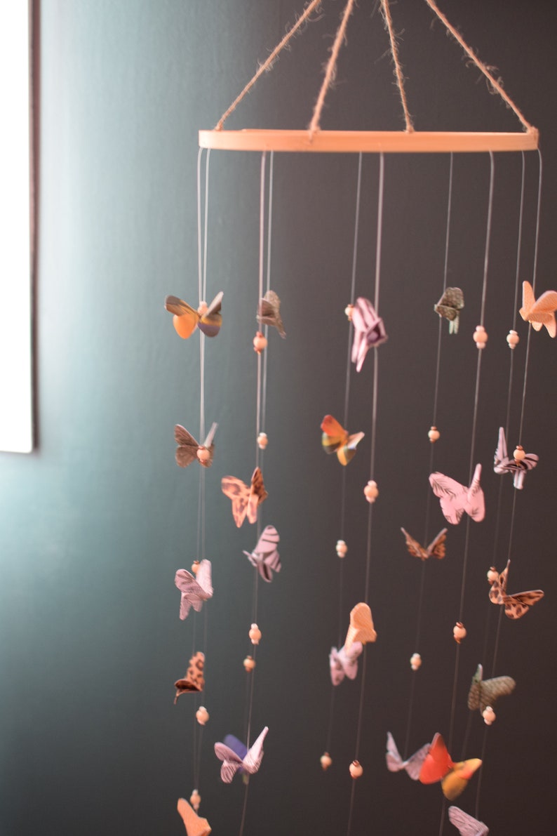 Puede incluir: Un m&oacute;vil de madera con un marco circular y m&uacute;ltiples cuerdas que cuelgan. Cada cuerda tiene un peque&ntilde;o adorno de papel en forma de mariposa de colores. Las mariposas est&aacute;n hechas de diferentes patrones y colores, incluyendo rosa, azul, verde y amarillo. El m&oacute;vil est&aacute; colgado del techo contra una pared verde oscuro.