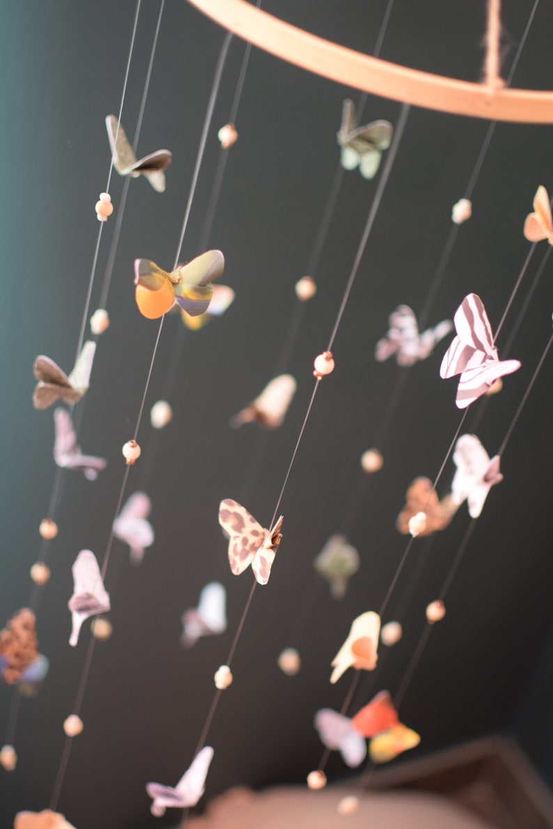 Puede incluir: Un m&oacute;vil con un aro de madera y varias mariposas de papel colgando de &eacute;l. Las mariposas son de varios colores y patrones, incluyendo rayas blancas y negras, estampado de leopardo y amarillo y verde.