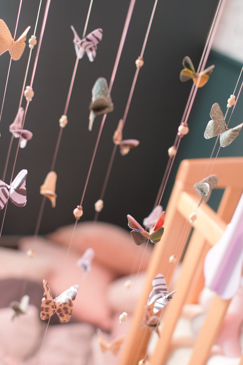 Puede incluir: Un m&oacute;vil con mariposas de papel de colores que cuelgan de cuerdas con cuentas de madera. Las mariposas son de varios patrones y colores, incluyendo naranja, verde, negro y blanco.