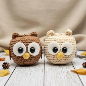 May include: Two crocheted owl-shaped keychains. One is brown, the other is cream-colored. Both have large white eyes with black pupils, yellow beaks, and small ear tufts. They are displayed on a white wooden surface with autumn leaves and pinecones.