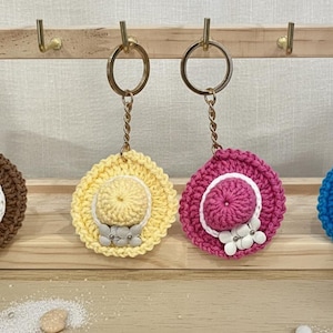 Può includere: Quattro portachiavi a forma di cappello all'uncinetto marroni, gialli, rosa e blu. Ogni cappello ha un bordo bianco e un fiore. Sono appesi a un supporto in legno con ganci dorati.