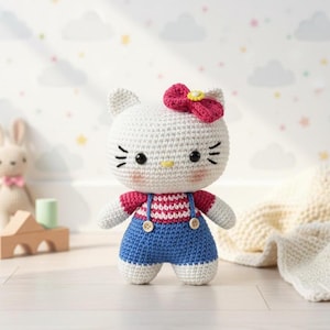 Puede incluir: Muñeca de ganchillo hecha a mano que se asemeja a un popular personaje de dibujos animados de un gato. La muñeca tiene una cabeza blanca con ojos negros, bigotes y un lazo rosa. Viste una camisa a rayas rojas y blancas con un peto azul.