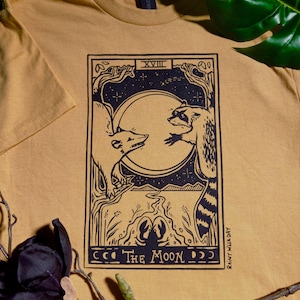 以下が含まれることがあります： マスタード色のTシャツ。白黒のタロットカードのデザインが特徴です。カードには、満月の前で向かい合うキツネとアライグマが描かれており、下部には「The Moon」の文字があります。「Rainy Weekday」の文字が右側に縦に印刷されています。