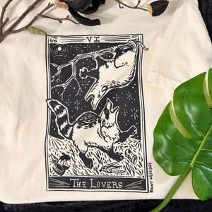 以下が含まれることがあります： オフホワイトのTシャツ。黒と白のタロットカードのデザインが特徴です。グラフィックは、枝からぶら下がっている動物と遠吠えしている動物の2つの動物の姿を描いています。「The Lovers」の文字が下部にあり、側面に「Rainy Week Day」と印刷されています。