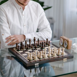 Może przedstawiać: Drewniany zestaw szachowy z ciemnobrązowymi i jasnymi figurami ułożonymi na szachownicy w szachownicę. Szachownica stoi na szklanym stole. Zegar szachowy jest widoczny w tle. Figury szachowe są wykonane z drewna.