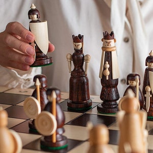 Może przedstawiać: Drewniany zestaw szachowy z ręcznie malowanymi figurami w stylu średniowiecznym. Figury są ustawione na czarno-białej szachownicy. Ręka sięga po konia.