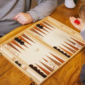 May include: A wooden backgammon board game with black and white checkers and dice. The game is set up on a light-colored surface with brown and white triangular points. Two red dice are held in a person's hand.