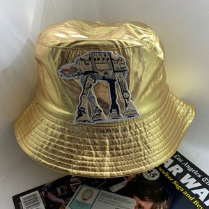 May include: A gold-coloured bucket hat with an embroidered AT-AT walker patch. The hat has a metallic sheen. A magazine with the title "Star Wars" is visible below the hat.