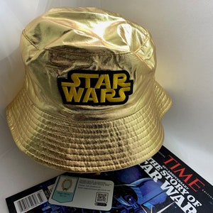 May include: Gold metallic bucket hat with the Star Wars logo patch. The hat is sitting on a white surface next to a TIME magazine with the headline "The Story of Star Wars".