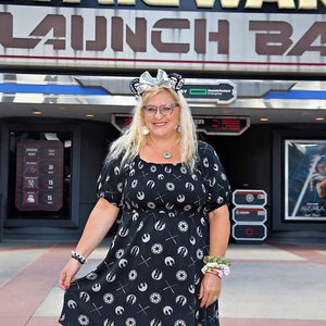 May include: A woman wearing a black and white dress with a Star Wars Rebel Alliance symbol pattern. She is standing in front of a Star Wars Launch Bay sign at Disney World. She is wearing a silver headband with mouse ears and glasses.