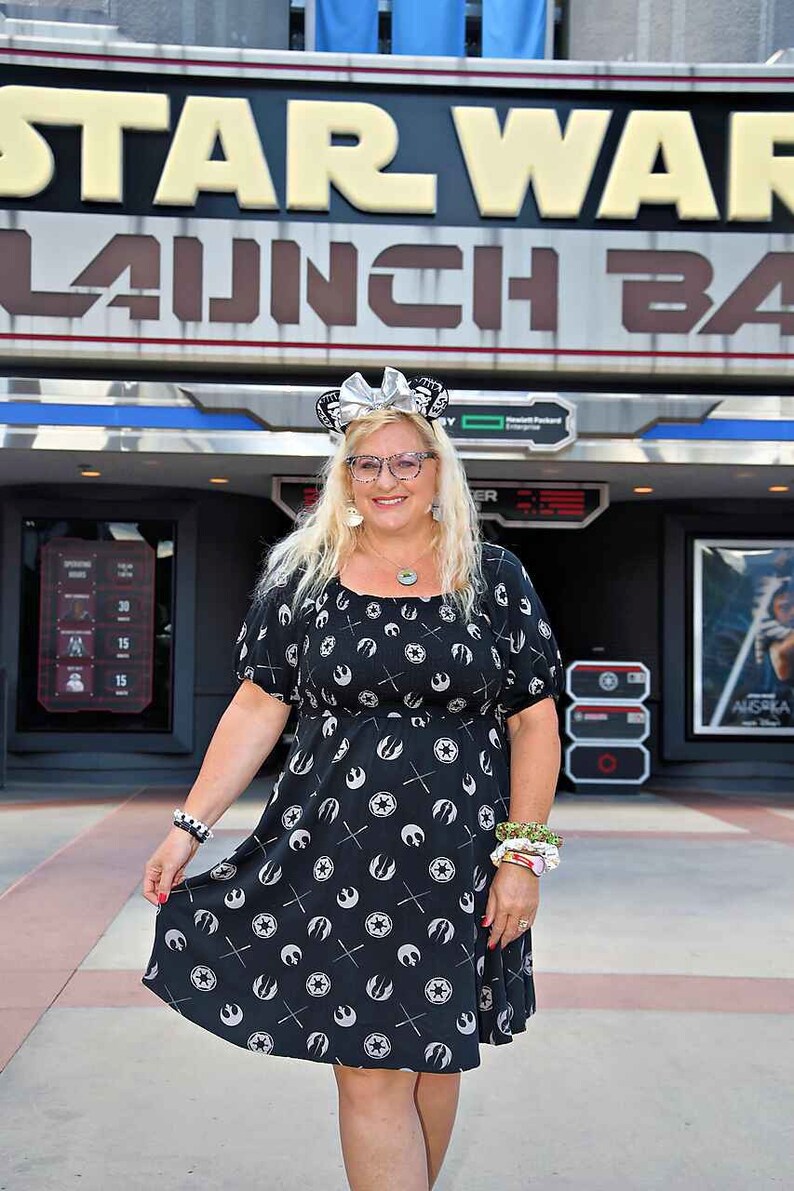 May include: A woman wearing a black and white dress with a Star Wars Rebel Alliance symbol pattern. She is standing in front of a Star Wars Launch Bay sign at Disney World. She is wearing a silver headband with mouse ears and glasses.