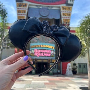 May include: Black Minnie Mouse ear headband with a large bow and sequined detailing. The ears are round and the bow is central. The headband is decorated with small, sparkling elements. The background shows a building with the text "Mickey & Minnie's Runaway Railway".