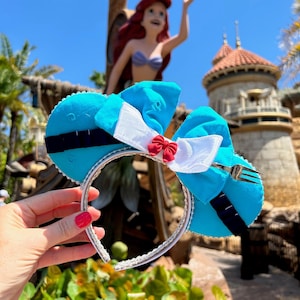 May include: A pair of light blue mouse ears with a white bow and a small red bow in the center. The ears have a black stripe and a silver headband. A fork is attached to the bow.