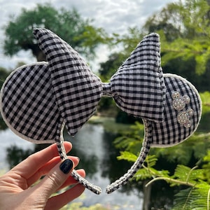 May include: A black and white gingham mouse ear headband with a matching bow. The ears have a white trim and are embellished with rhinestone Mickey Mouse silhouettes. The headband is held up by a hand in front of a blurred outdoor background.