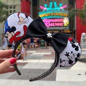 May include: Mickey Mouse ear headband with a black bow and a small white flower accent. The ears feature Mickey Mouse in a sorcerer's outfit and a black and white Mickey Mouse design. The headband is black with black beading.