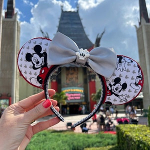 May include: Mickey Mouse ear headband with a grey bow and a white crown accent. The ears feature a white background with Mickey Mouse illustrations and a red trim. The headband is black with a grey braided design.