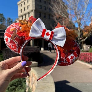 May include: Red and white Minnie Mouse ears with a white bow and a Canadian flag. The ears are decorated with maple leaves, Canadian flags, and a bear.