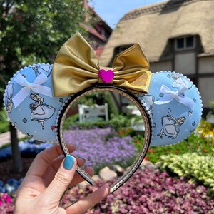 May include: A headband featuring two light blue ears decorated with a repeating pattern of a character in a dress, accented by small white bows on each ear. A large metallic gold bow sits at the centre, adorned with a small, bright pink heart charm.