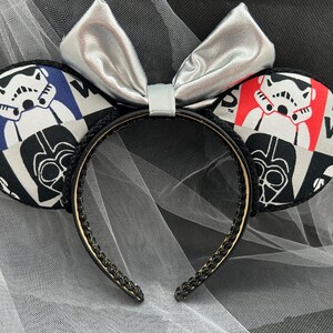 May include: Black and white Star Wars themed Minnie Mouse ears with a silver bow. The ears feature a patchwork design with images of Darth Vader and Stormtroopers.