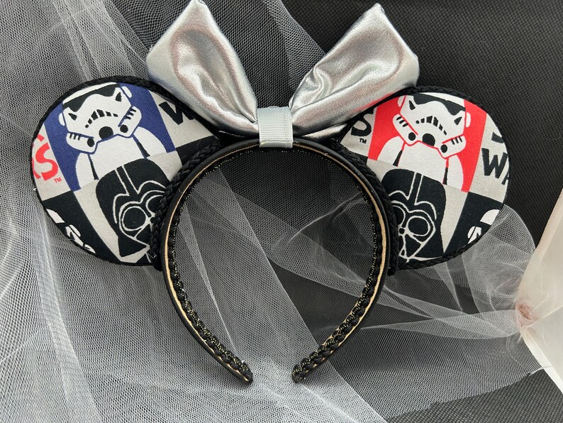 May include: Black and white Star Wars themed Minnie Mouse ears with a silver bow. The ears feature a patchwork design with images of Darth Vader and Stormtroopers.