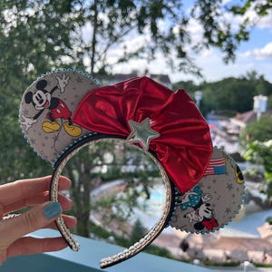 May include: Mickey Mouse ear headband with gray fabric ears featuring Mickey and Minnie Mouse, a red bow, and a silver star. The headband has a silver braided trim and a black interior.
