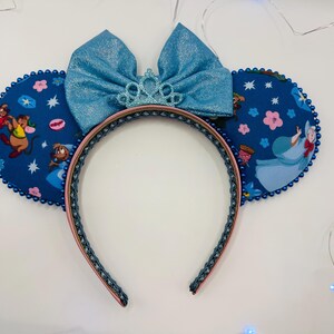 May include: A pair of mouse ears with a blue bow and a small tiara. The ears are navy blue with a pattern of Cinderella characters and pink flowers. The headband is navy blue with a pink trim.