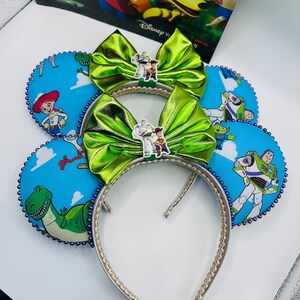 May include: Two headbands featuring blue fabric ears with a cloud pattern and Toy Story character prints including Woody, Buzz Lightyear, and Rex. Each headband has a metallic lime green bow centred with a small Woody and Buzz Lightyear charm.