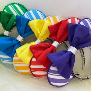 May include: A collection of colorful headbands with large fabric bows and striped ear accents. The bows are in shades of green, blue, yellow, red, and purple. The ear accents have white stripes. The headbands have silver trim.