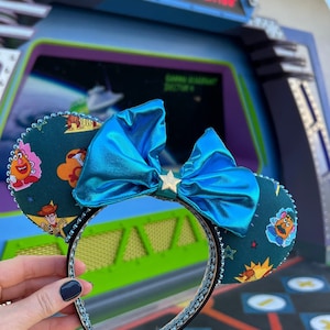 May include: Dark teal mouse ears headband with a large, shiny blue bow and a gold star. The ears are decorated with Toy Story characters. The headband is trimmed with silver rhinestones. The background shows a Space Ranger station.