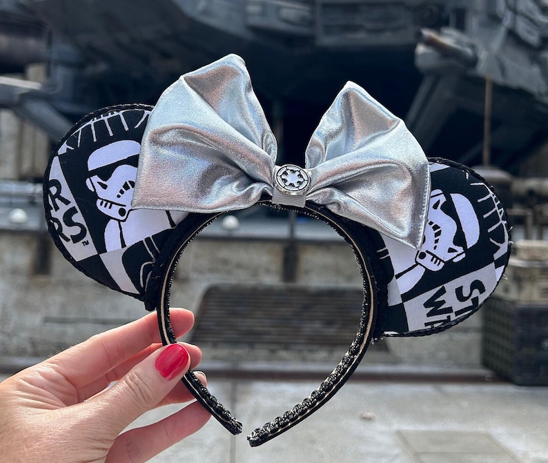 May include: Black and white Star Wars themed mouse ears with a silver bow. The ears feature a Stormtrooper design with the text "Star Wars" on one ear and "Rebel Alliance" on the other.