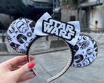 May include: White and black Star Wars themed Minnie Mouse ears with a white bow that says "STAR WARS" in black letters. The ears are covered in black and white Star Wars character silhouettes.