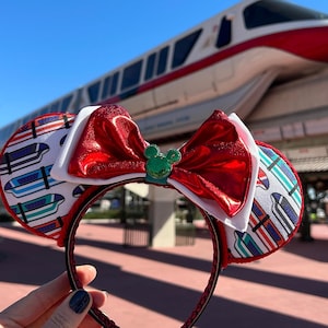 May include: Red and white Minnie Mouse ear headband with a large red bow and a green Mickey Mouse emblem. The ears feature a pattern of colorful train cars. The headband is held up in front of a monorail.