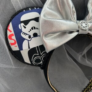 May include: Black and white Star Wars themed mouse ears with a silver bow and a black headband. The ears feature a Stormtrooper and Darth Vader.