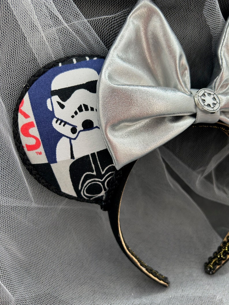 May include: Black and white Star Wars themed mouse ears with a silver bow and a black headband. The ears feature a Stormtrooper and Darth Vader.