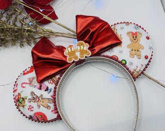 May include: A headband featuring two circular ears with a white fabric cover printed with gingerbread characters, candy canes, and holiday treats. A large, shiny metallic red bow sits between the ears, adorned with a small gingerbread couple charm.
