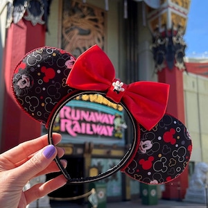 May include: Mickey Mouse ear headband in black and red, featuring a large red bow and a small white flower. The ears display a Mickey Mouse pattern, and the text "RUNAWAY RAILWAY" is seen through the center.