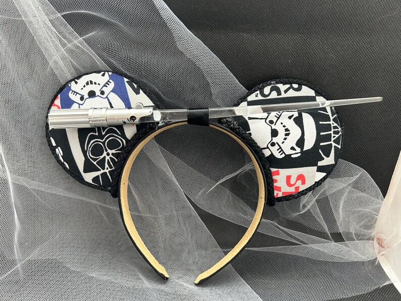 May include: Black and white Star Wars themed mouse ears with a silver lightsaber accessory. The ears are made of fabric and have a black headband.
