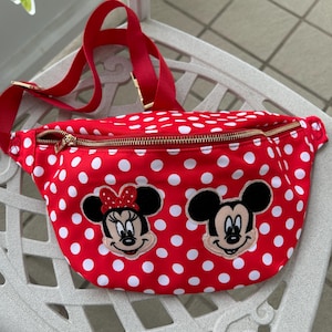 May include: Red fanny pack with white polka dots and a gold zipper. The bag features embroidered images of Mickey and Minnie Mouse. The adjustable strap is red.