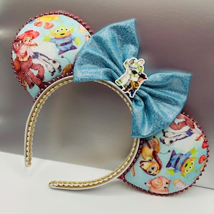 May include: Headband featuring two circular ears with a light blue fabric printed with Toy Story characters including Woody, Jessie, and an alien. The ears are trimmed with small red beads. A large, shimmering blue bow sits between the ears.