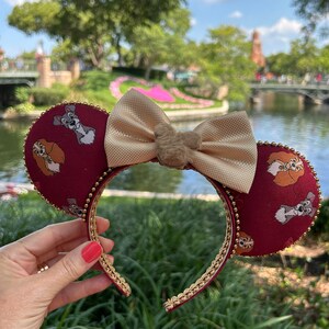 May include: Burgundy mouse ears with a gold trim and a beige bow. The ears feature a repeating pattern of cartoon dogs. The bow has a small, fluffy accent in the centre. The headband is held by a person.
