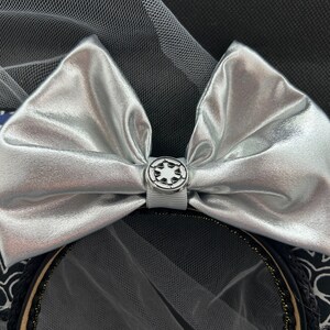 May include: Silver bow with a white and silver Imperial symbol button, on a black headband with a white and black Stormtrooper design.